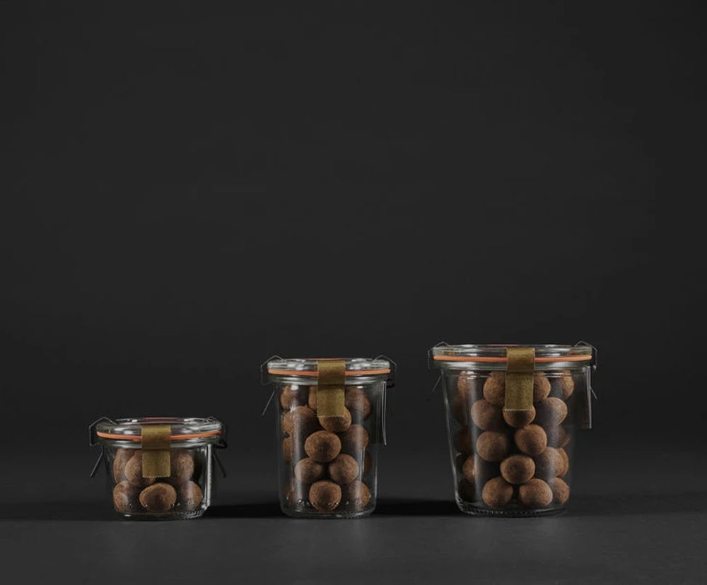Three clear glass jars with varying sizes on a dark background, filled with chocolate covered caramelized hazelnuts with bourbon sea salt covered in cocoa powder and coffee. each jar is sealed with metal clips with a hanging tag attached underneath the right clip.