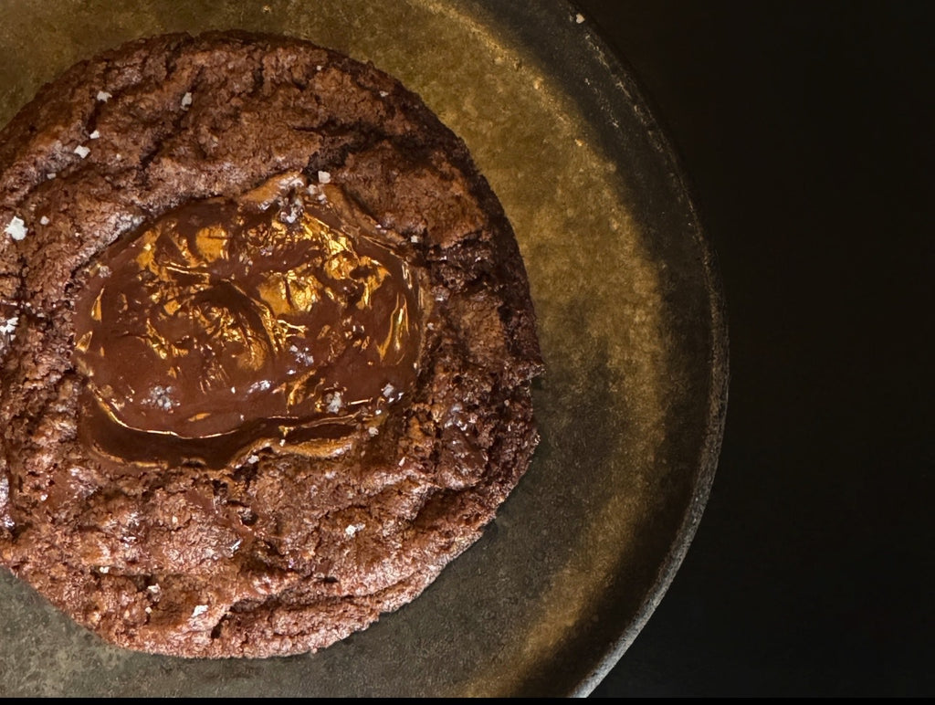 A dark chocolate cookie with a melted chocolate filling in the center and sprinkled with salt on a dark round stoneware dish.